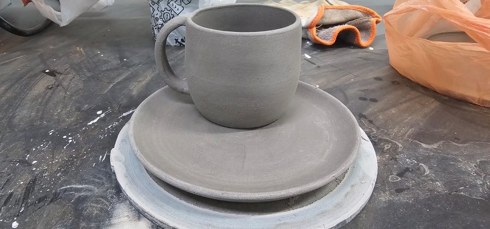 Caneca e pratos ainda sem esmalte apoiados sobre uma bancada de trabalho.