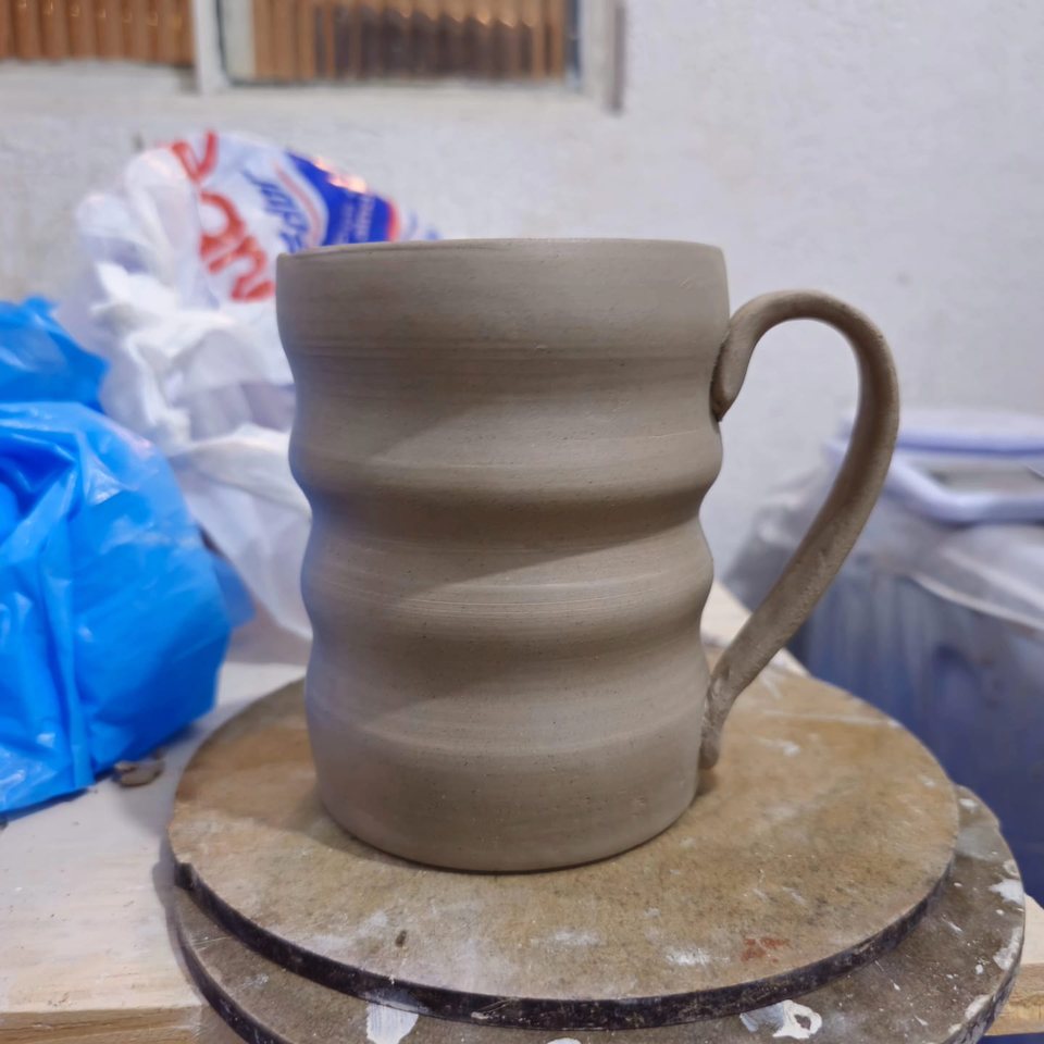 Caneca de argila crua com corpo ondulado e alça curva sobre o torno.